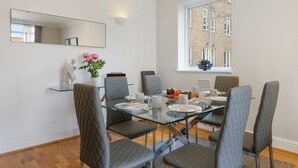 A dining area with a glass table, grey upholstered chairs, a white statue, and a window with a view of a brick building.