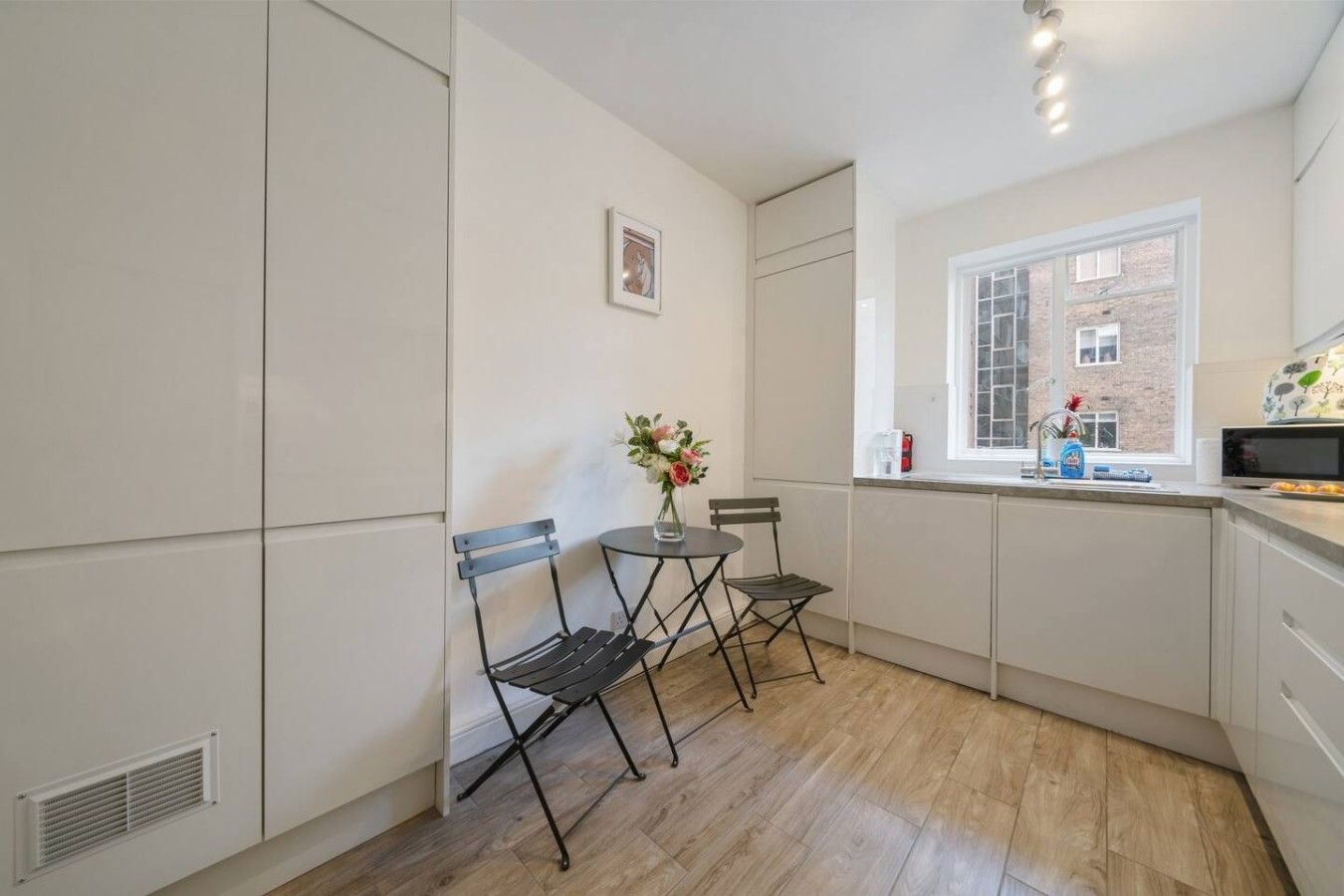 Een moderne keuken met witte kasten, een raam met uitzicht op een gebouw van baksteen, en een klein eethoekje met twee stoelen en een tafel.