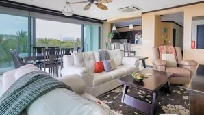 A modern living room with a sofa, armchairs, and a coffee table. There is a dining area with chairs and a table. The room has large windows and a ceiling fan. - Diamante del sol 601N (Jacó)