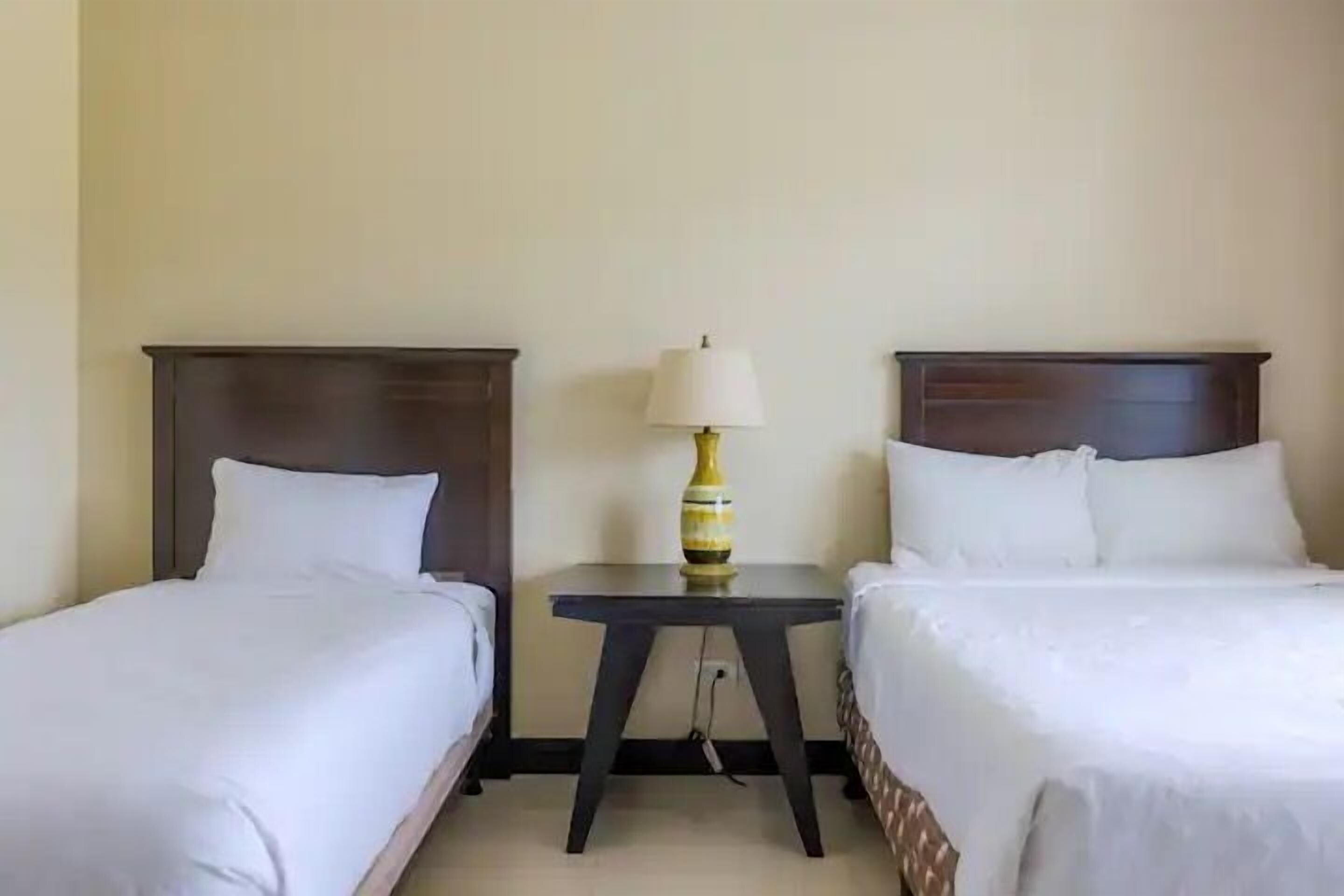Een hotelkamer met twee eenpersoonsbedden, een tafeltje met een lamp en een effen muur.