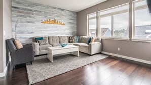 A modern living room with a grey sofa, a white coffee table, and a grey armchair. There is a decorative wall piece above the sofa and a large window on the right side. - Majestic Views with a Hot Tub! (Seattle)