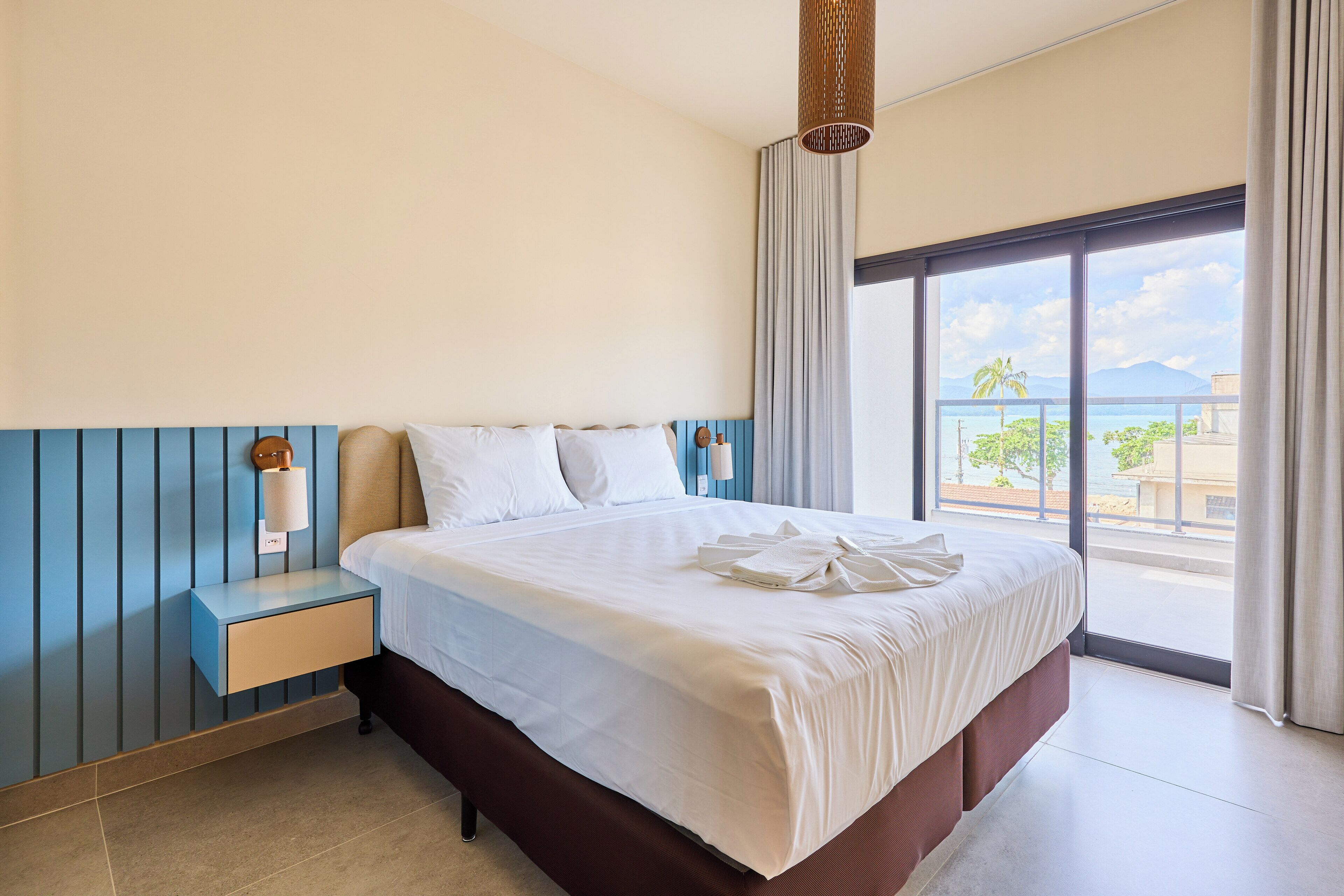 A hotel room with a large bed, a nightstand, a lamp, and a view of the ocean through a large window.