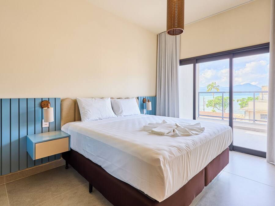 A hotel room with a large bed, a nightstand, a lamp, and a view of the ocean through a large window.