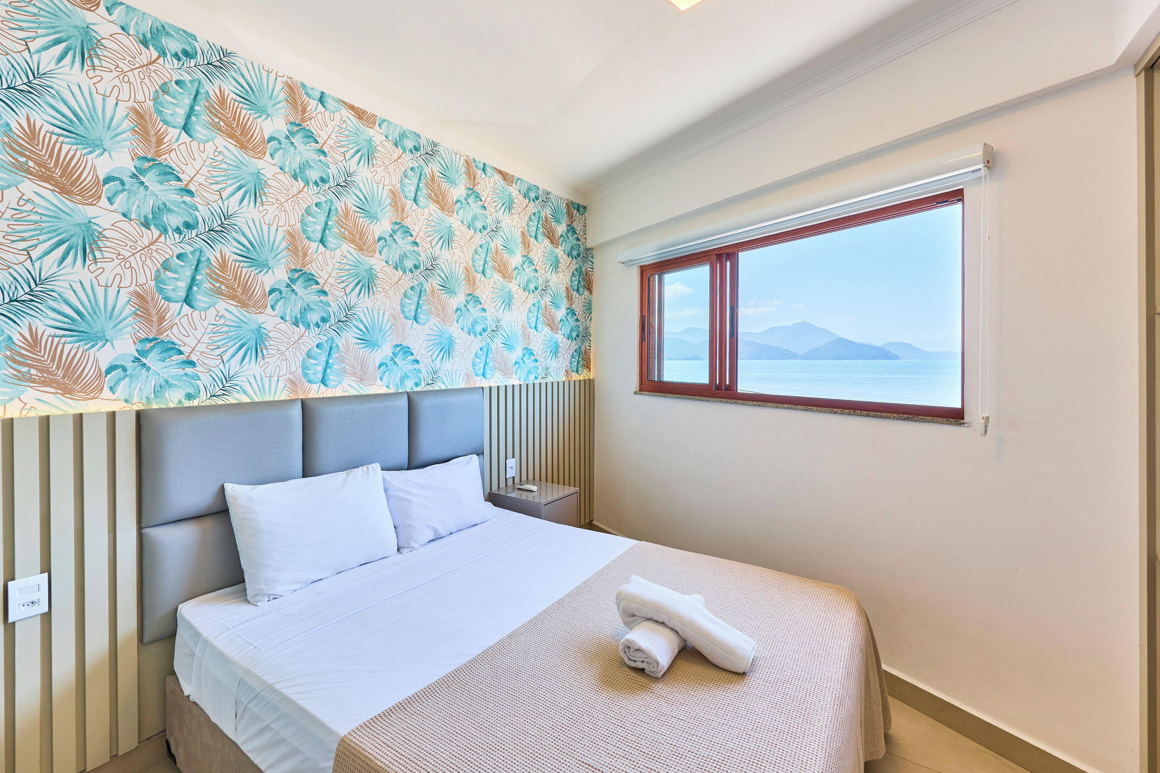 Una cama bien tendida con sábanas blancas, una pared con diseño y una ventana con vistas al mar.