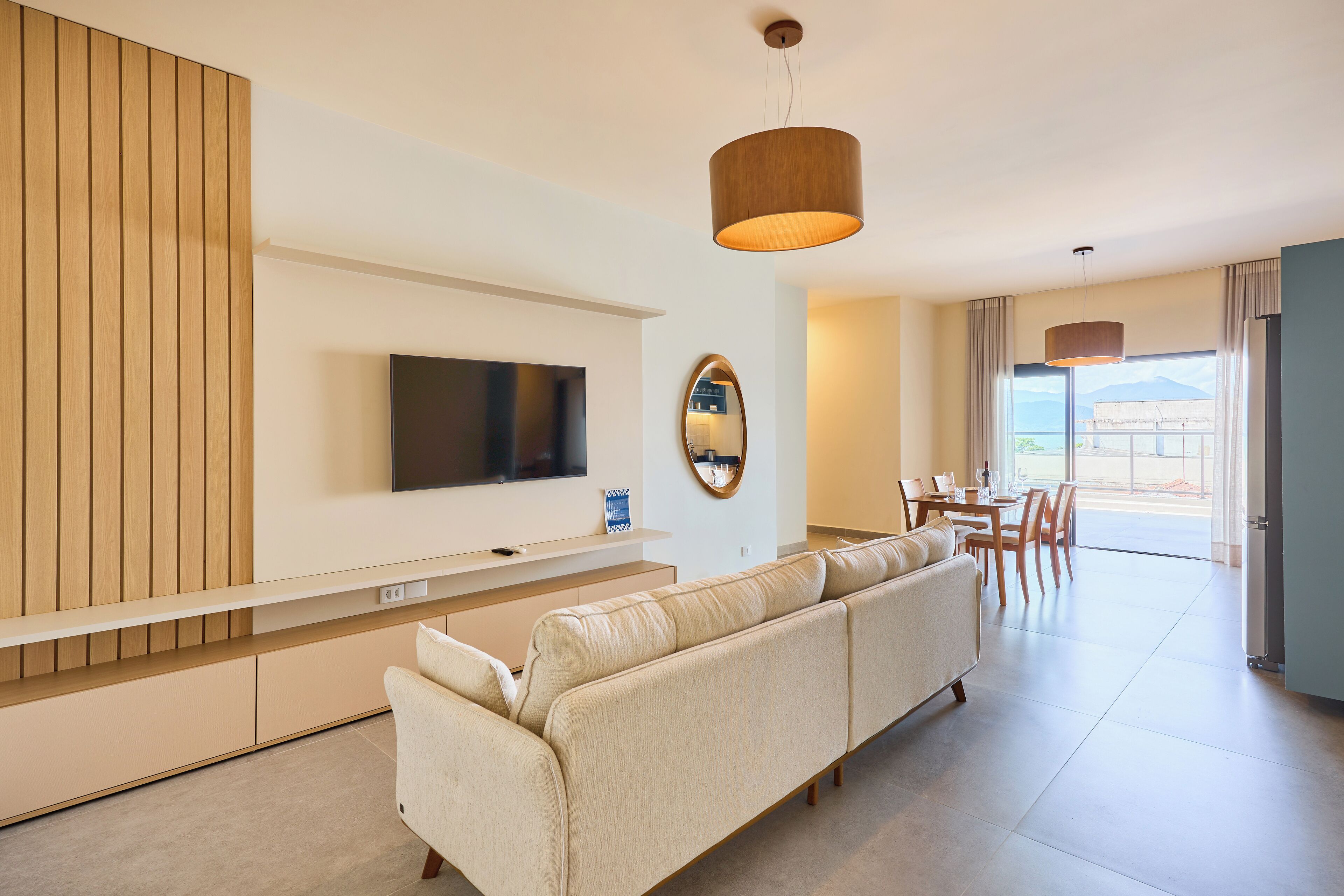 A modern living room with a large flat-screen TV, a sofa, a dining area with chairs, and a view of the outdoors.