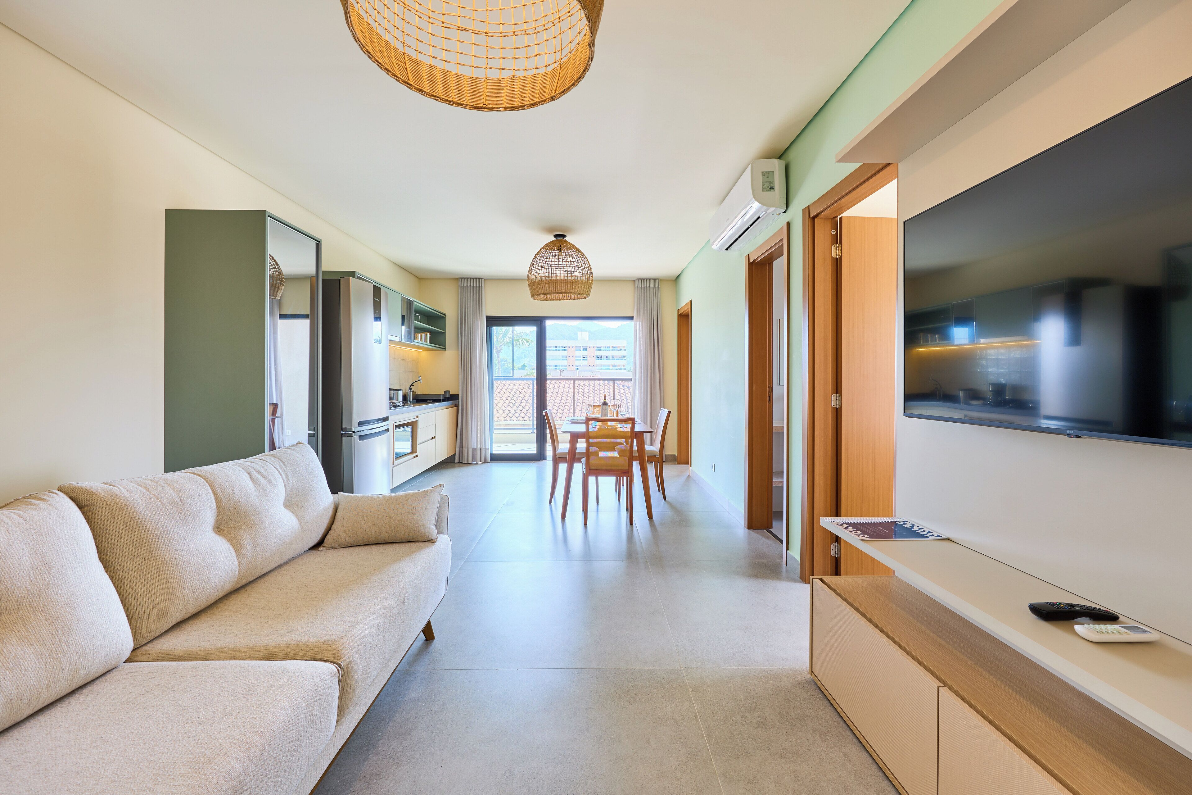 A modern living room with a sofa, a dining table with chairs, a TV, and a kitchen area with cabinets and a window.