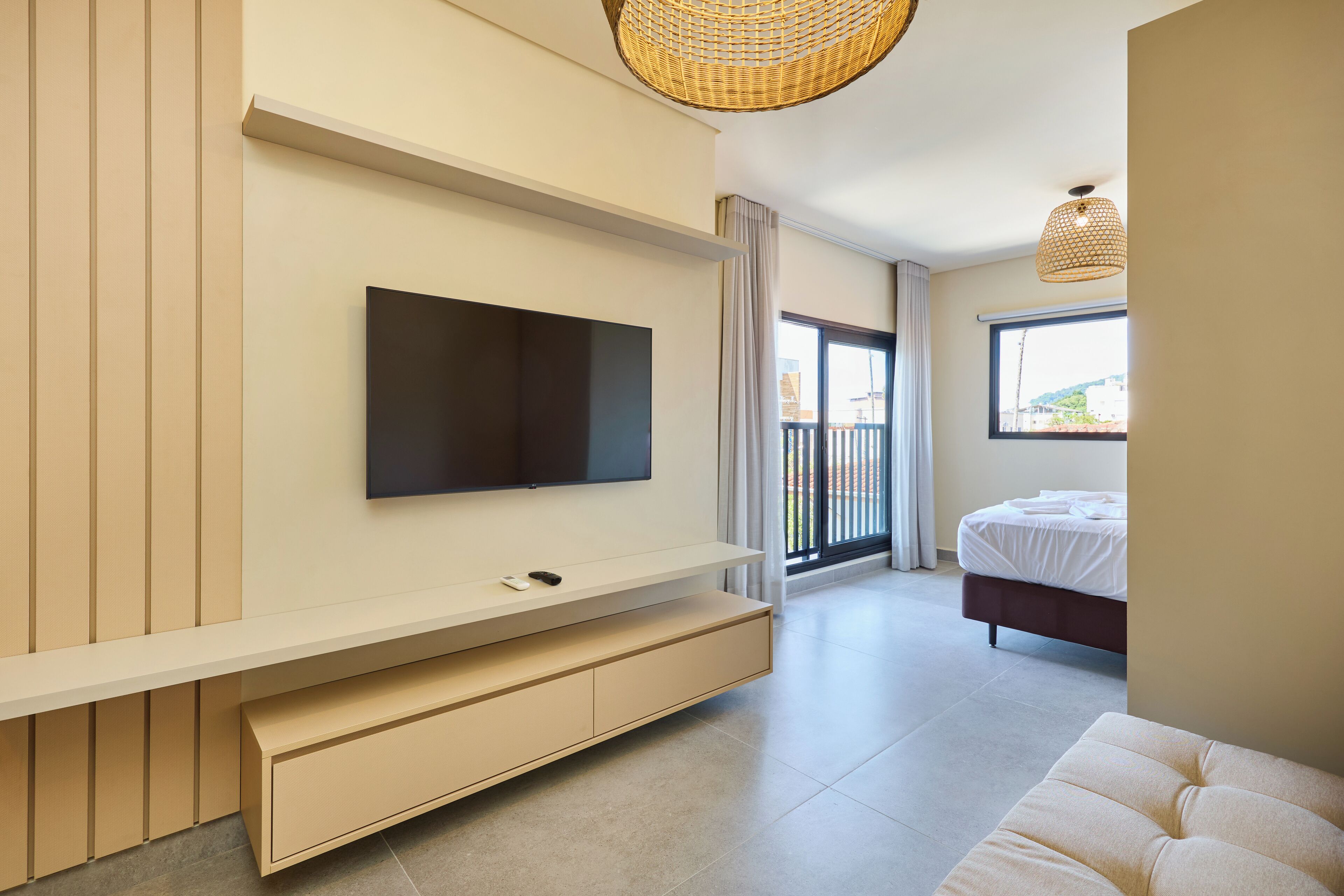 A modern bedroom with a large TV mounted on the wall, a bed with white linens, and a balcony with a view.