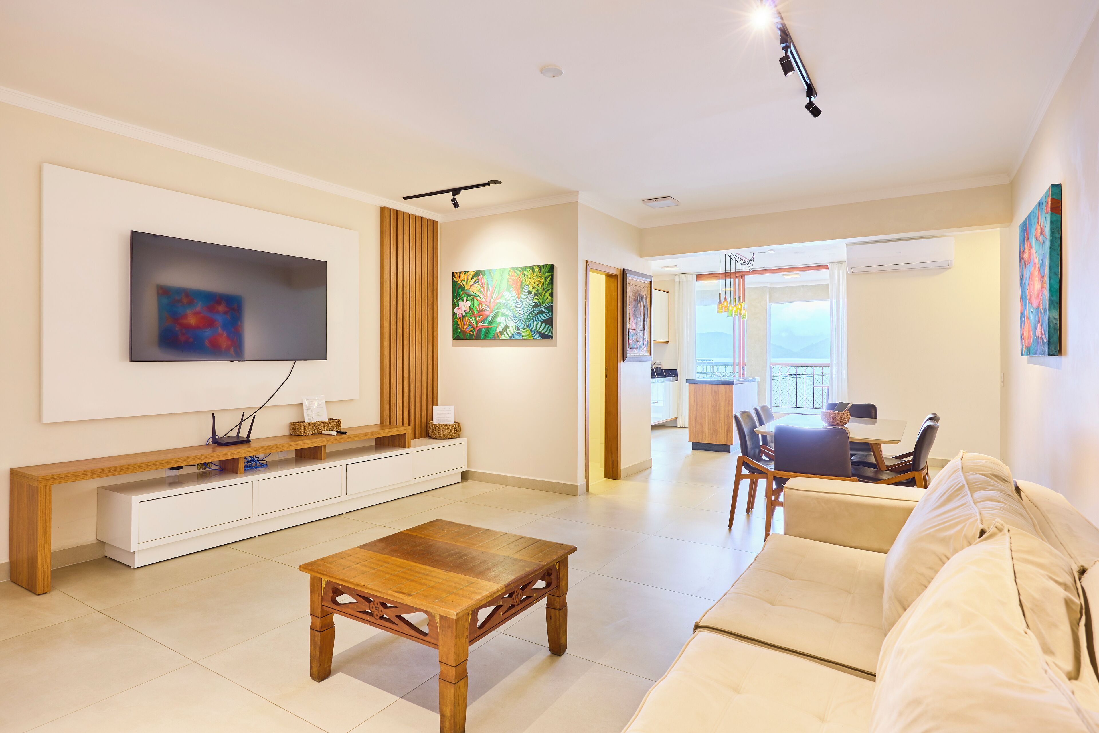 A modern living room with a wooden coffee table, a beige sofa, a TV mounted on the wall, and a colorful painting.