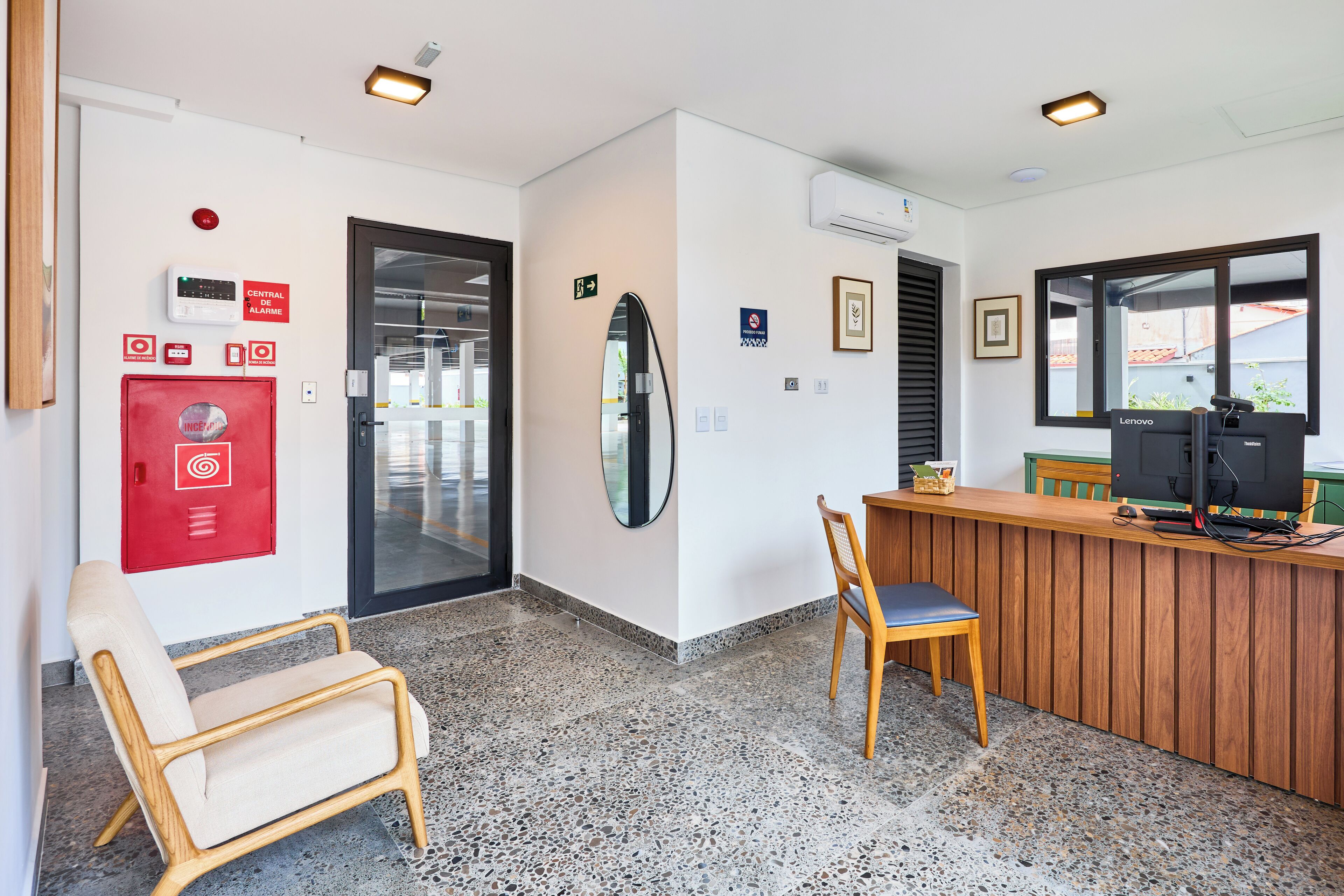 A reception area with a wooden desk, a computer, and a chair. There is a red fire alarm box on the wall and a door leading to another room.