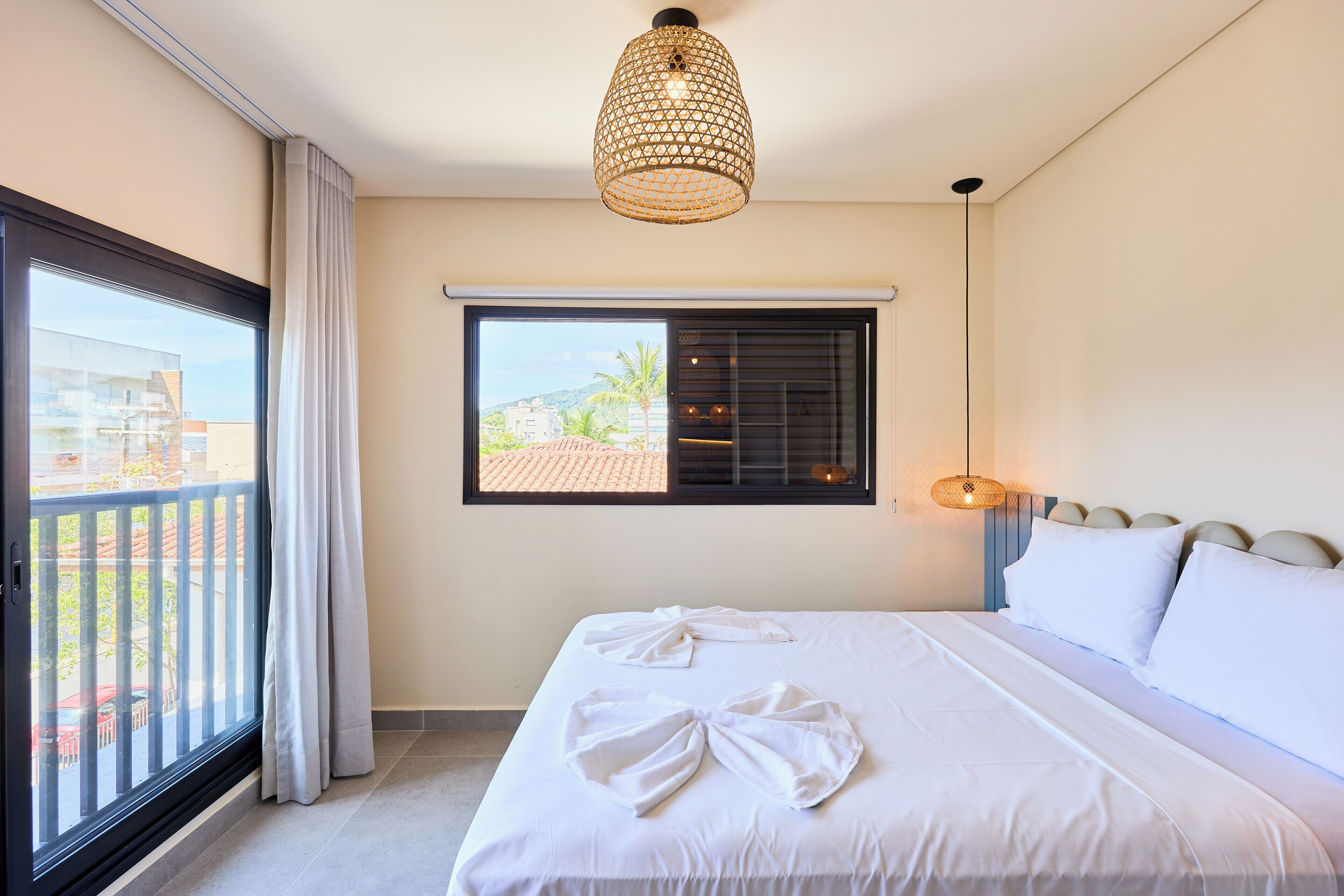 A hotel room with a large bed, a window with blinds, a balcony, and a hanging light fixture.