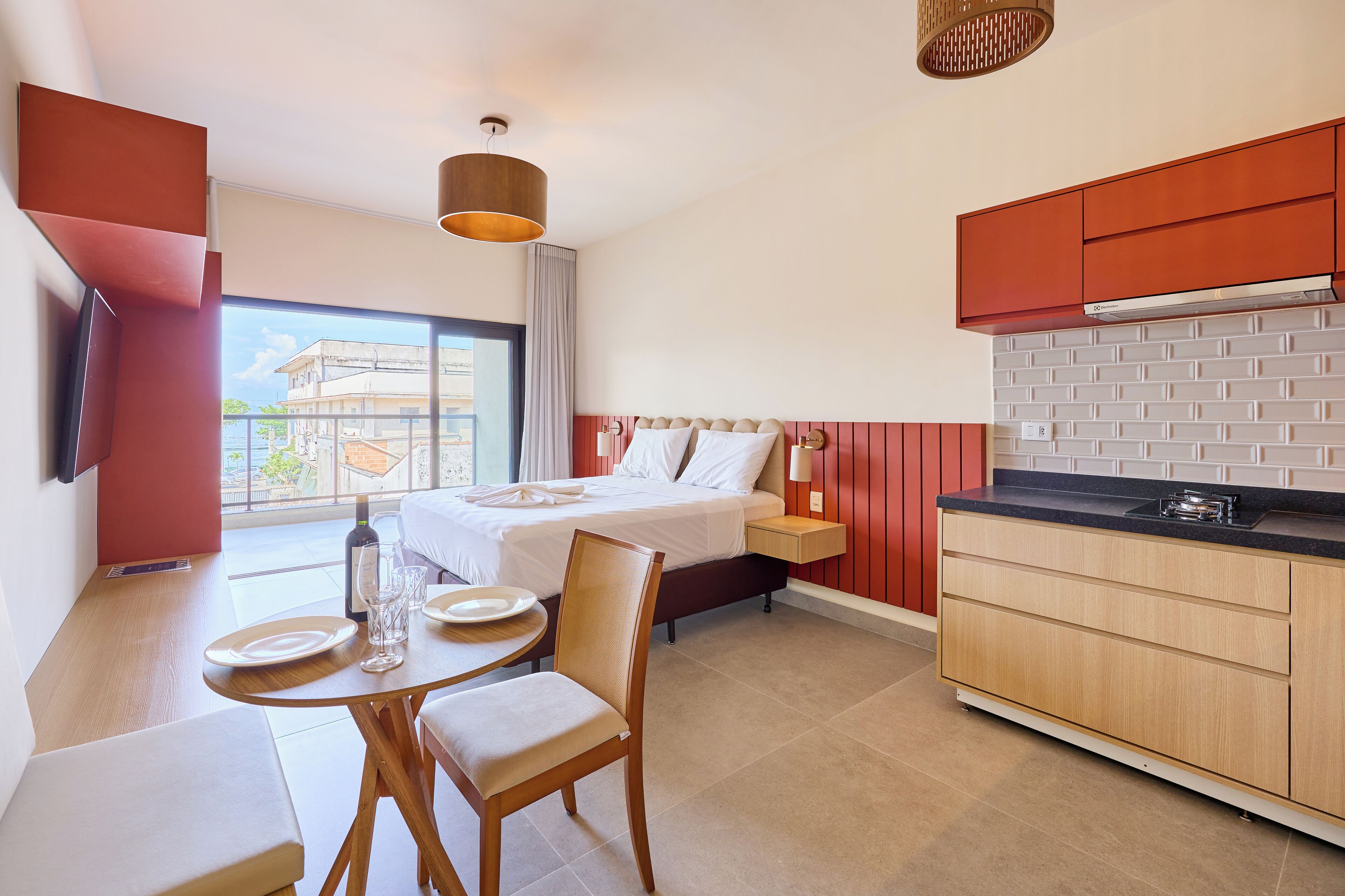 Ett hotellrum med en säng, ett matbord med stolar, ett köksutrymme och ett stort fönster med utsikt över byggnader.