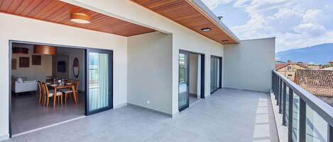 Een ruim balkon met houten plafond, glazen deuren naar een eethoek en uitzicht op het omliggende landschap.