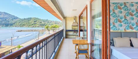 A balcony with a wooden table and chairs, a bed with a patterned headboard, and a view of a beach and mountains.