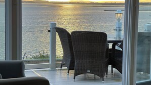 Un balcon avec des fauteuils en osier et une table, donnant sur une étendue d’eau où le soleil se couche.