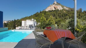 Uma área à beira da piscina com mesa e cadeiras, vista para uma colina com uma estátua, e um edifício com antenas parabólicas.