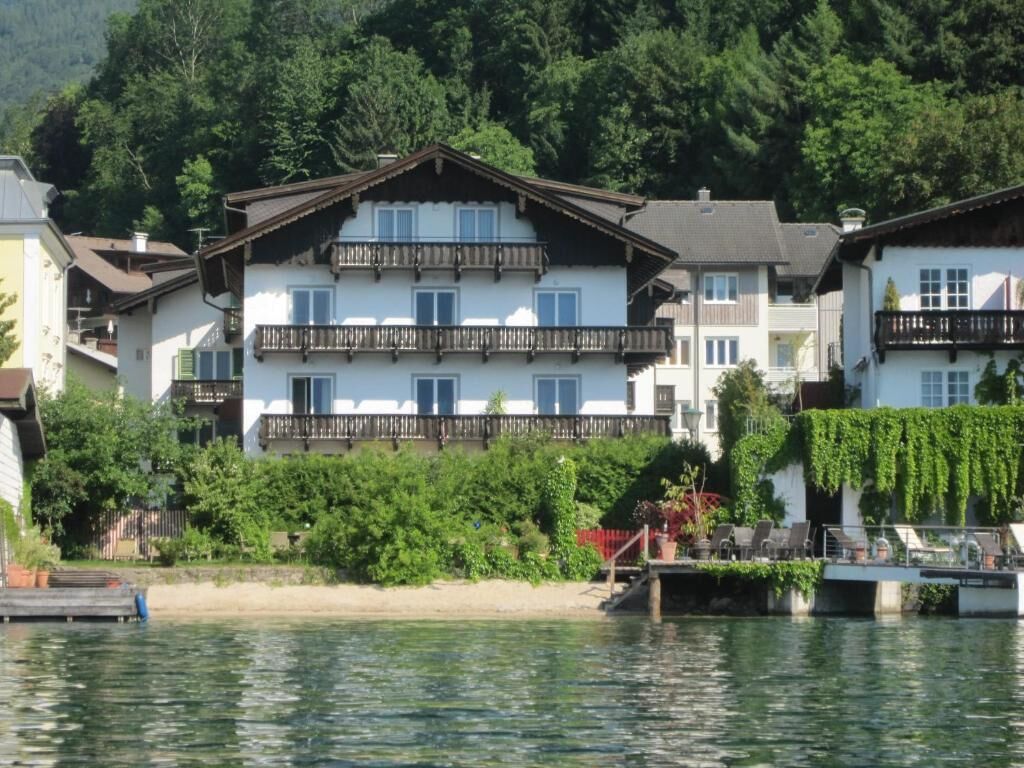 Ein mehrstöckiges Gebäude mit Balkonen, an einem Gewässer mit Anlegesteg und Grünflächen im Hintergrund.