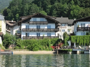Un edificio de varios pisos con balcones, ubicado junto a una masa de agua con un muelle y vegetación al fondo.