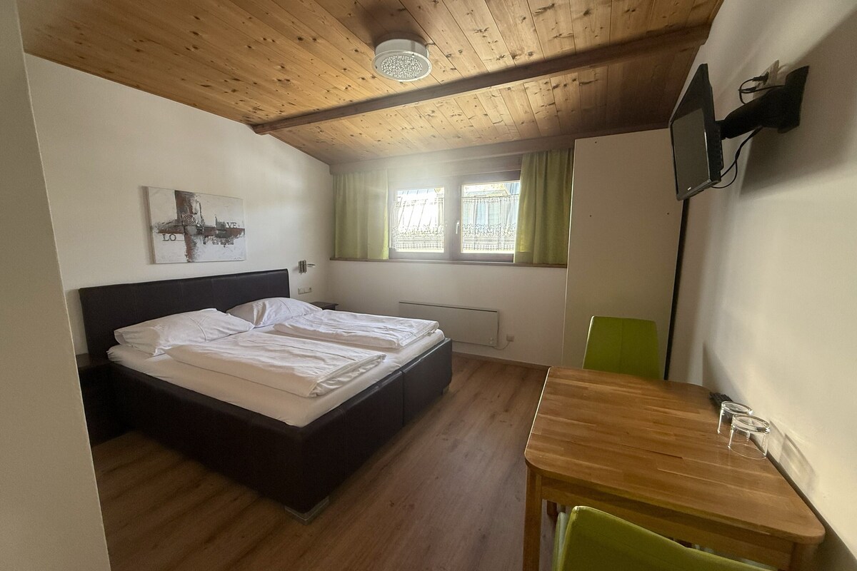 Una habitación con cama, una mesa, una silla, ventana con cortinas, radiador y televisión fijada a la pared.