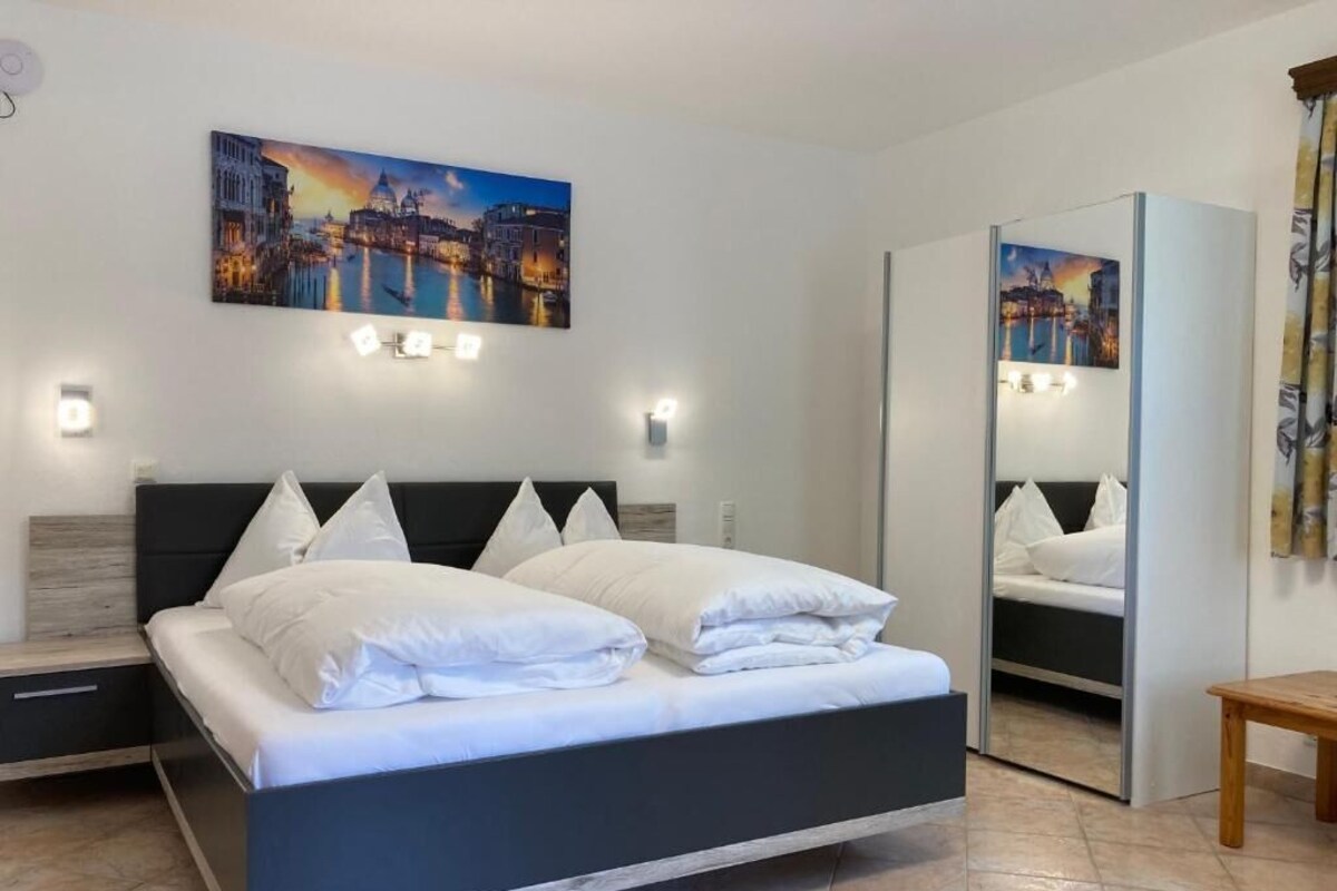 Un dormitorio moderno con cama, mesitas de noche, un espejo y una pintura de paisaje urbano en la pared.