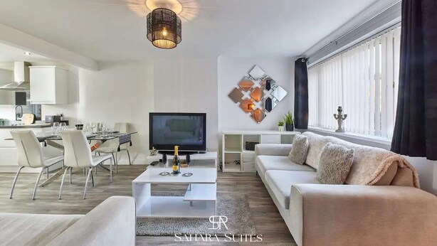 Ein modernes Wohnzimmer mit einem Flachbildfernseher, weißen Sofas, einer Essecke mit Tisch und Stühlen sowie einer Küche im Hintergrund.