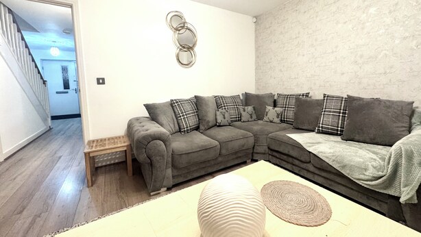 Una sala de estar con un sofá seccional gris, varios cojines, un jarrón blanco redondo y un mantel tejido sobre una mesa de color claro.