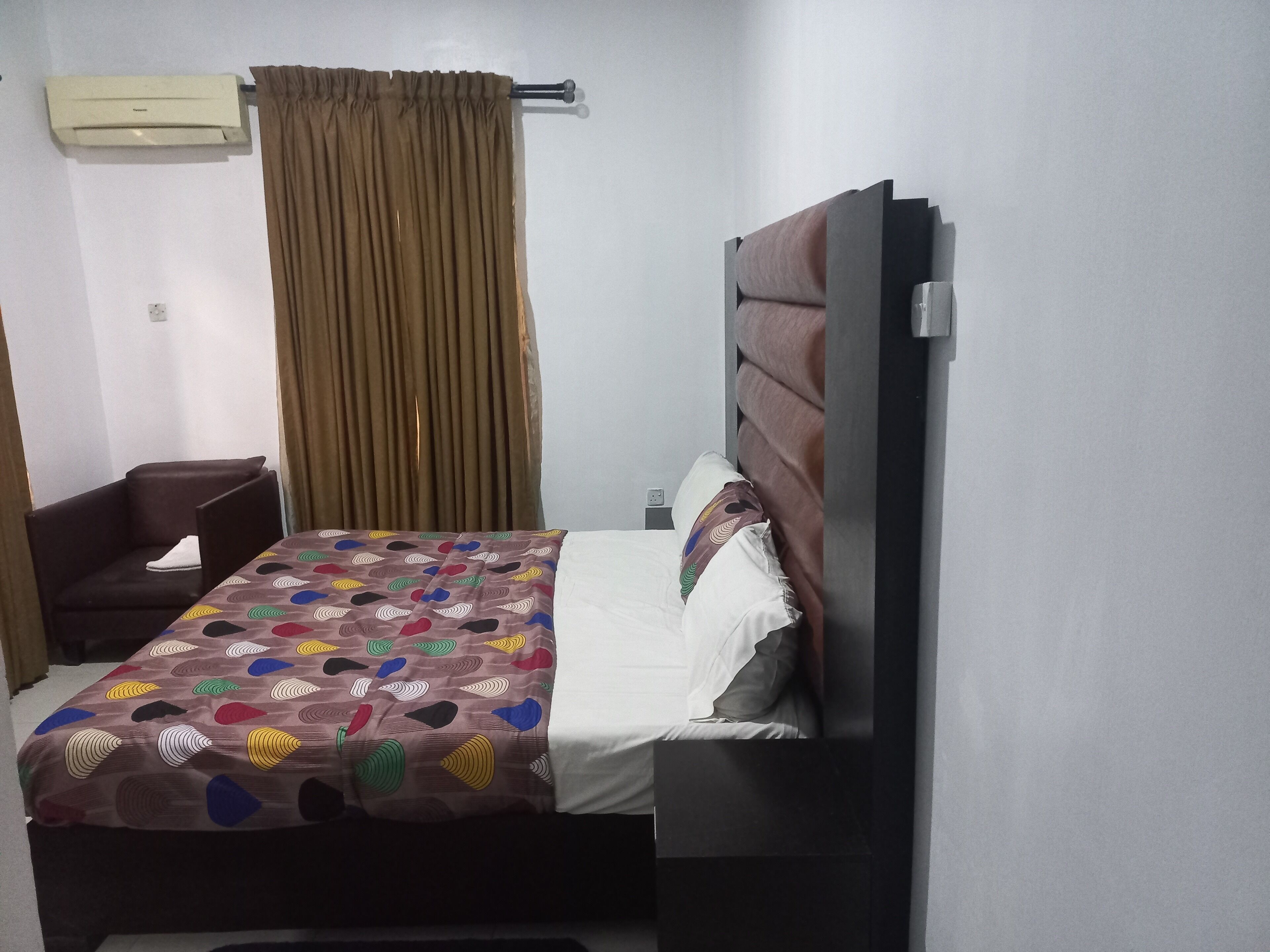 A hotel room with a bed, a chair, a desk, an air conditioning unit, and a window with curtains.