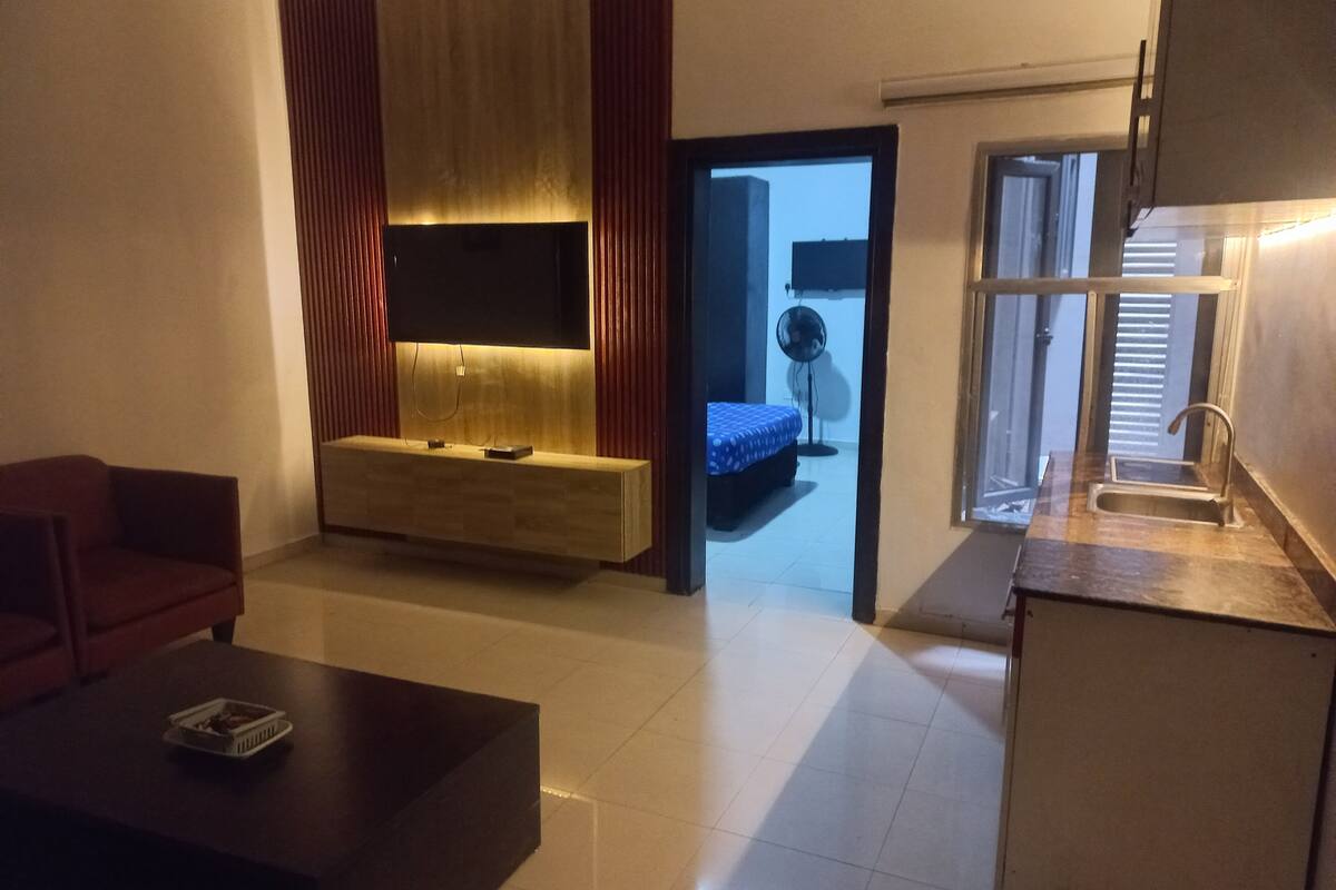 A modern living room with a flat-screen TV mounted on a wooden console, a red sofa, a coffee table, and a kitchen area with a sink.