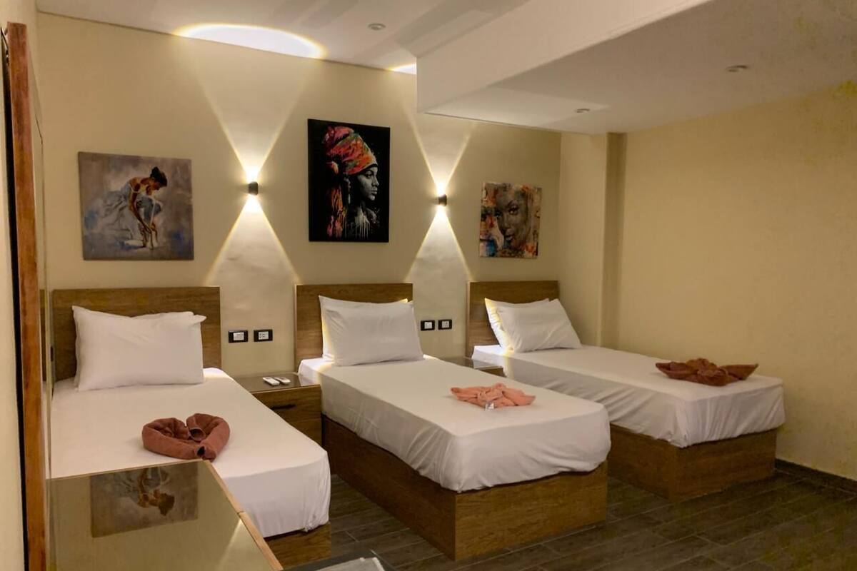 A hotel room with three single beds, a wooden headboard, white bedding, and artwork on the walls.