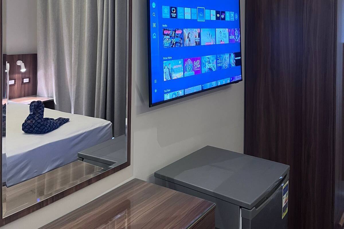 A hotel room with a large flat-screen TV mounted on the wall displaying a streaming service interface, a wooden dresser, a small refrigerator, and a mirror reflecting the room's interior.