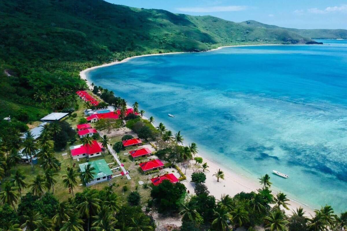 Vista aérea de un resort costero con playa, edificios de techo rojo y exuberante vegetación.