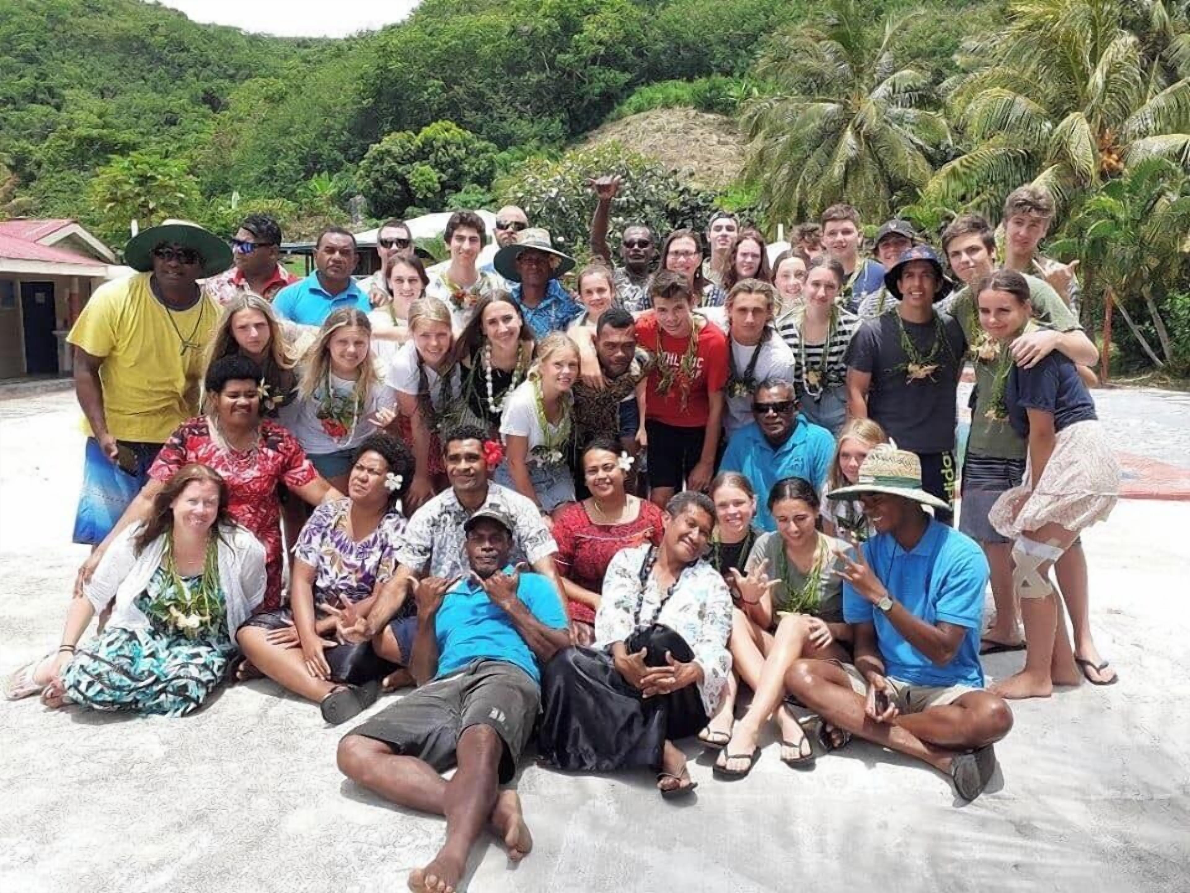 A group of people gathered outdoors, possibly at a beach or resort area with palm trees and a building in the background.