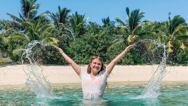 Iemand in een helderblauw zwembad met armen omhoog, omringd door palmbomen en een zandstrand.