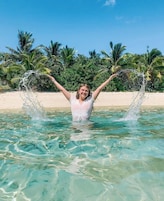 Una persona en una piscina azul cristalina con los brazos levantados, rodeada de palmeras y una playa de arena.