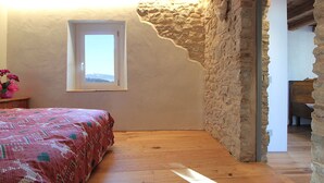 A bedroom with a wooden floor, a bed with a patterned bedspread, a window with a view, and stone walls.