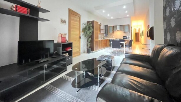 A modern living room with a black leather sofa, a glass coffee table, and a flat-screen TV. There is a kitchen area in the background with wooden cabinets and a dining table. - Bugibba Central Modern Apartment, Charming 2-bedroom apartment with AC,  WiFi  (San Pawl il-Baħar)