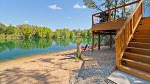 A wooden deck with a staircase leading to a covered area, overlooking a calm lake with trees in the background. - The Fairy House | 3 Beds 2 Baths (Elgin)