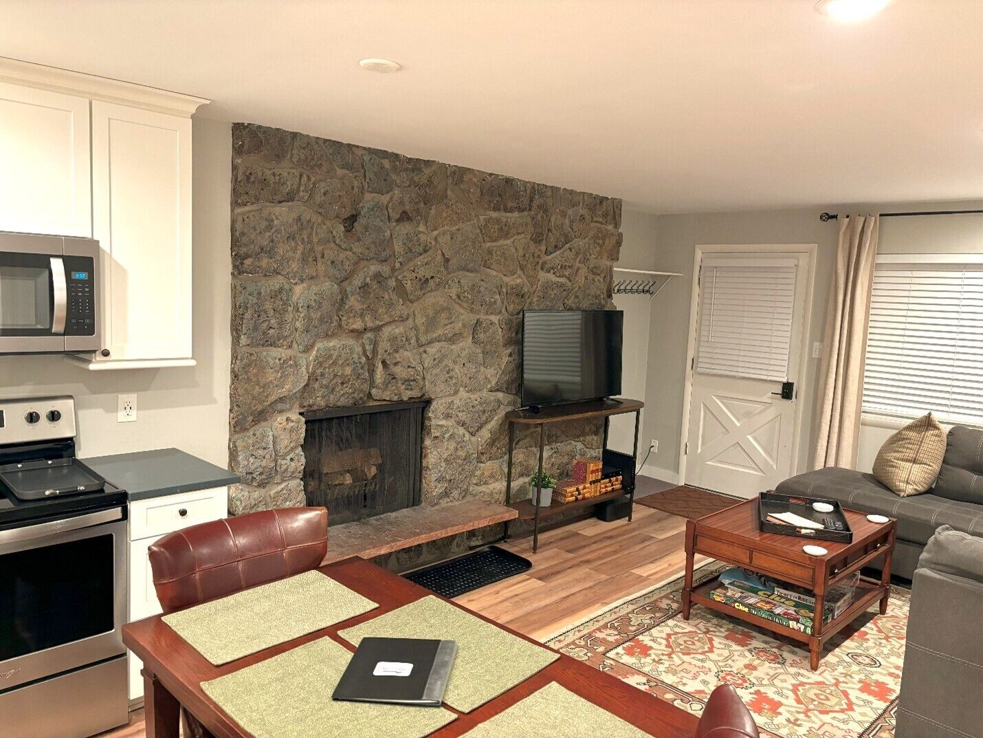 A modern living room with a stone fireplace, a television on a stand, a dining area with a table and chairs, and a kitchen with appliances.