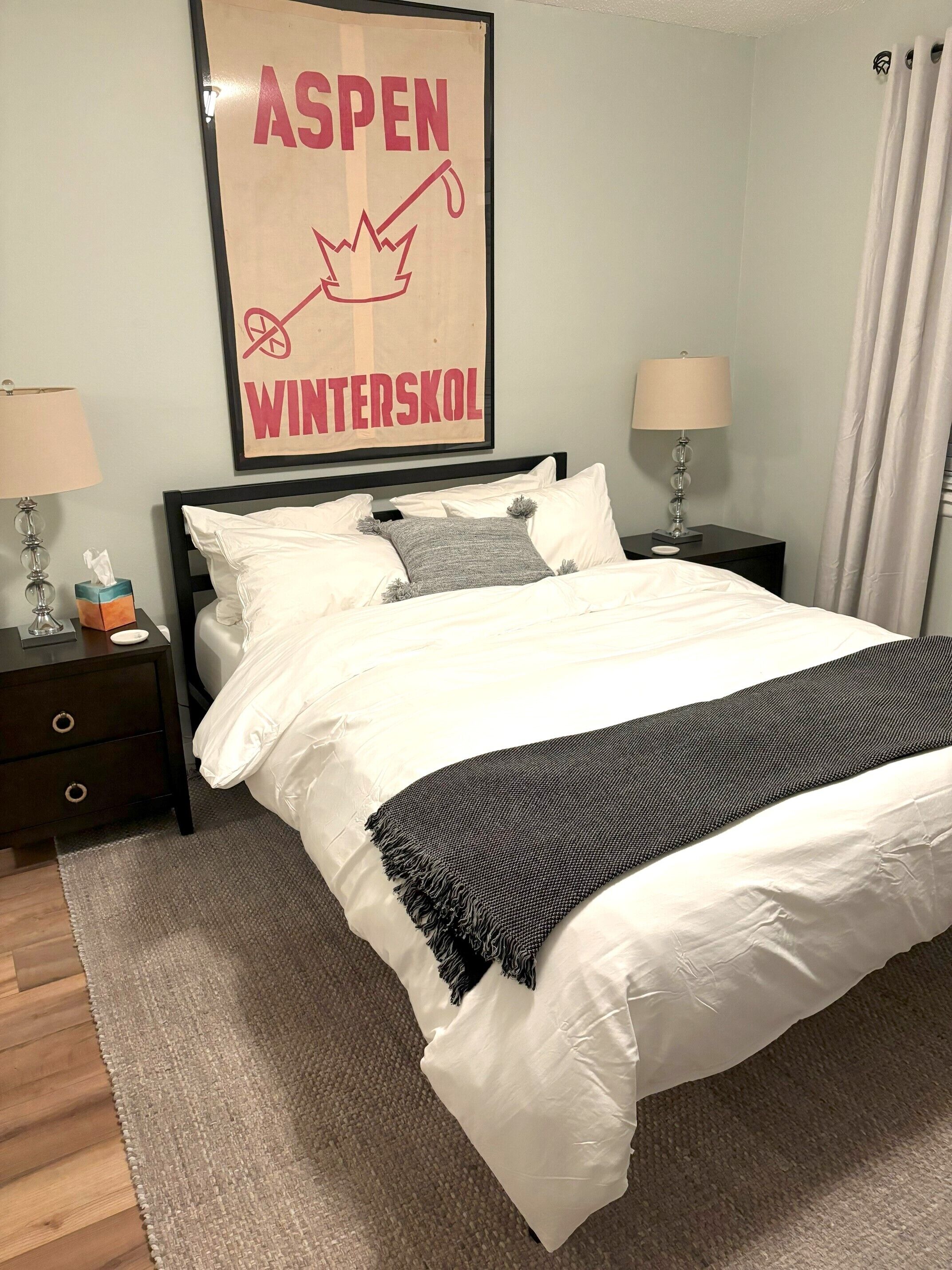 A neatly made bed with white and gray bedding, two bedside tables with lamps, and a framed sign above the bed.