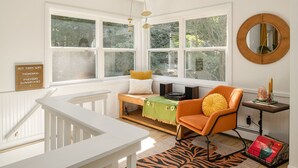 Une pièce lumineuse avec une grande fenêtre, un banc en bois, une chaise orange, un miroir rond et une petite table sur laquelle se trouve une bougie.