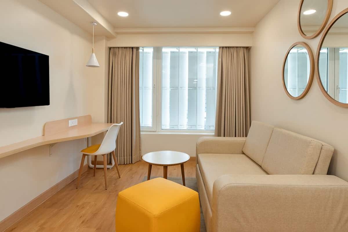 A modern living room with a sofa, a yellow ottoman, a small round table, a TV mounted on the wall, and a window with curtains.