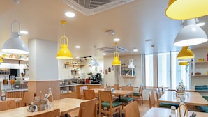A modern restaurant with wooden tables and chairs, yellow pendant lights, and a kitchen area in the background. - Bloom Hotel SG Highway (Ahmedabad)