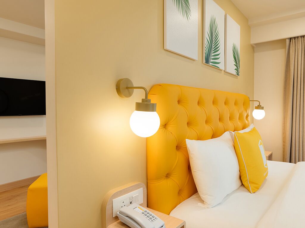 Een hotelkamer met een gestoffeerd geel hoofdeinde, wit beddengoed en een telefoon op het nachtkastje.
