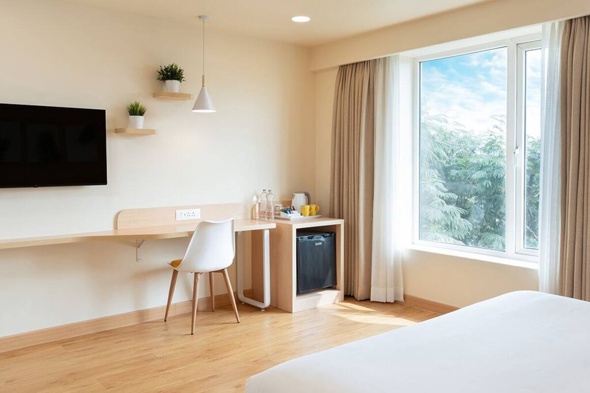 A modern hotel room with a bed, a desk with a chair, a TV mounted on the wall, and a window with curtains.