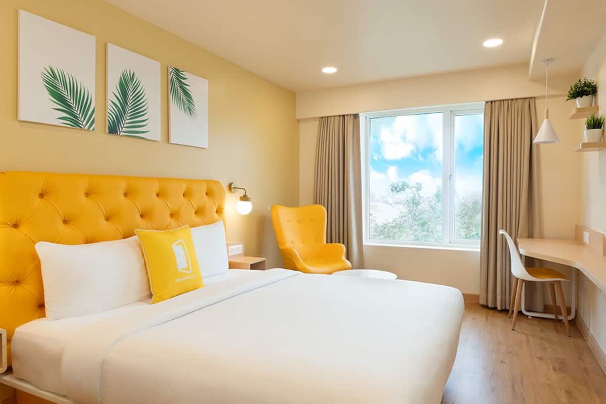 A hotel room with a large bed, a desk, two chairs, a window with curtains, and three palm leaf wall art pieces.