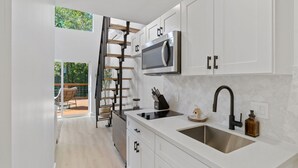 A modern kitchen with white cabinets, a stainless steel sink, and a built-in microwave. A wooden ladder leads to a balcony with a chair and a table. - The Bird House | 6 Beds 2 Baths (Bastrop County)