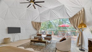 A spacious tented room with a large window, a flat-screen TV, a sofa, a coffee table, and a lamp. - The Bird House | 6 Beds 2 Baths (Bastrop County)