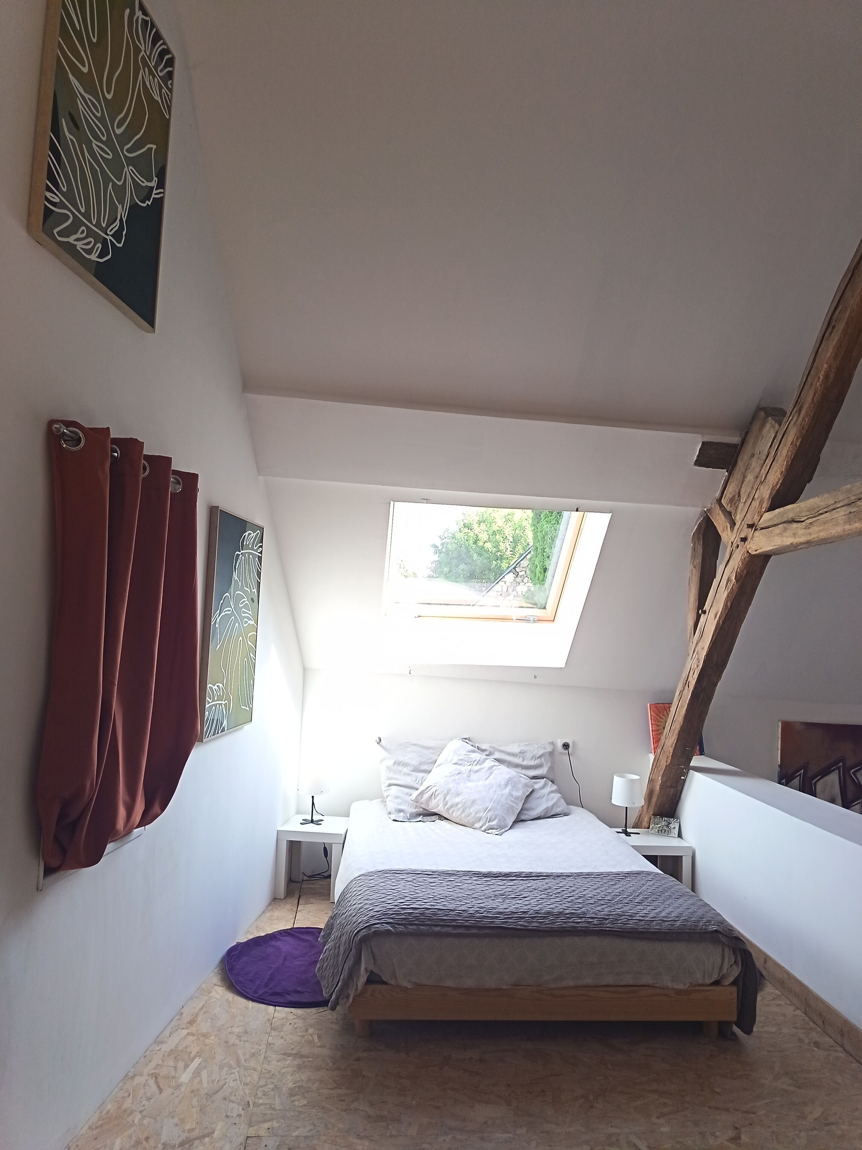 A bedroom with a sloped ceiling, a skylight, a bed with pillows, a nightstand, and a purple rug.