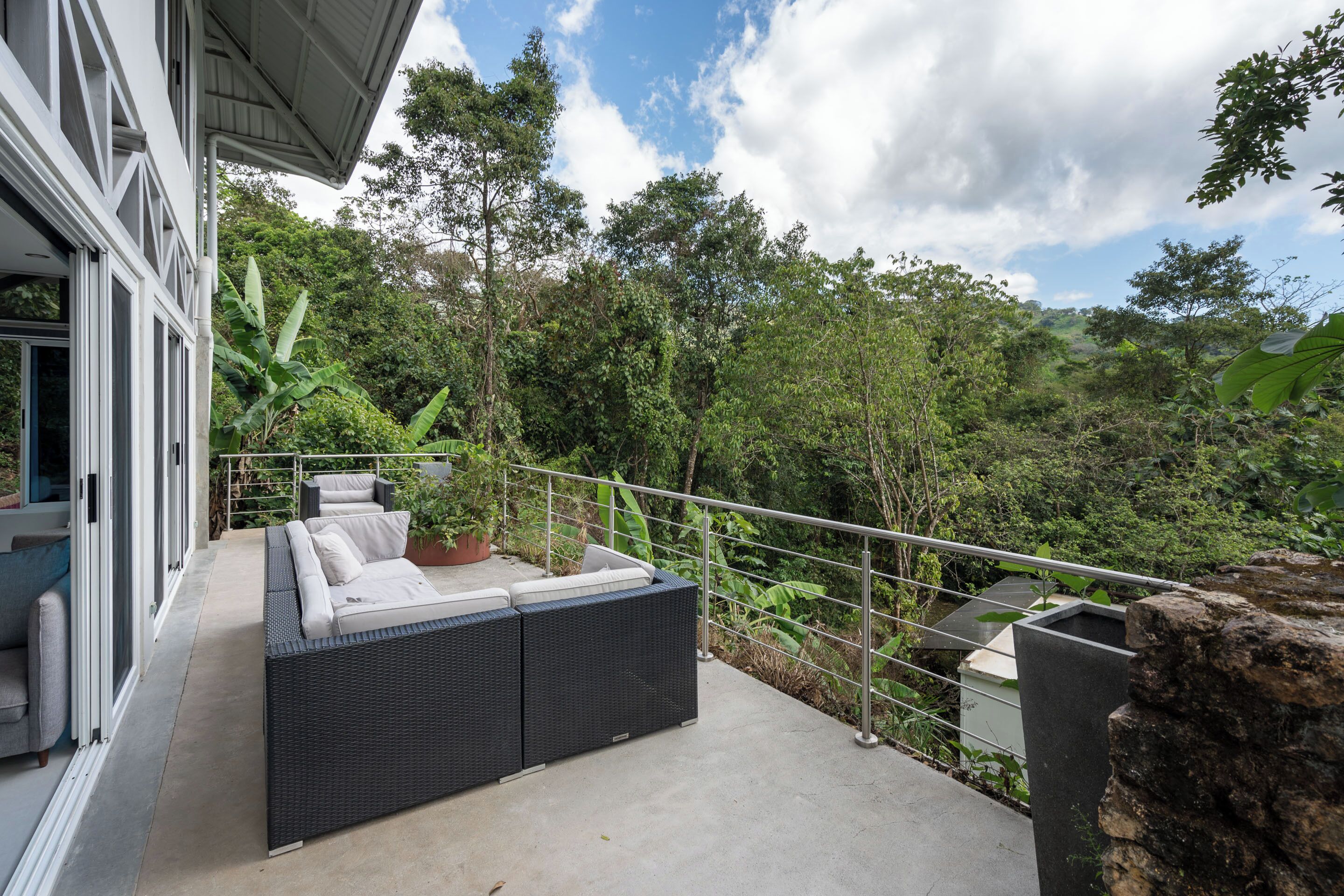 Ein Balkon mit einer schwarzen Rattan-Couch, einem Topfpflanzen und Blick auf eine üppige grüne Landschaft.