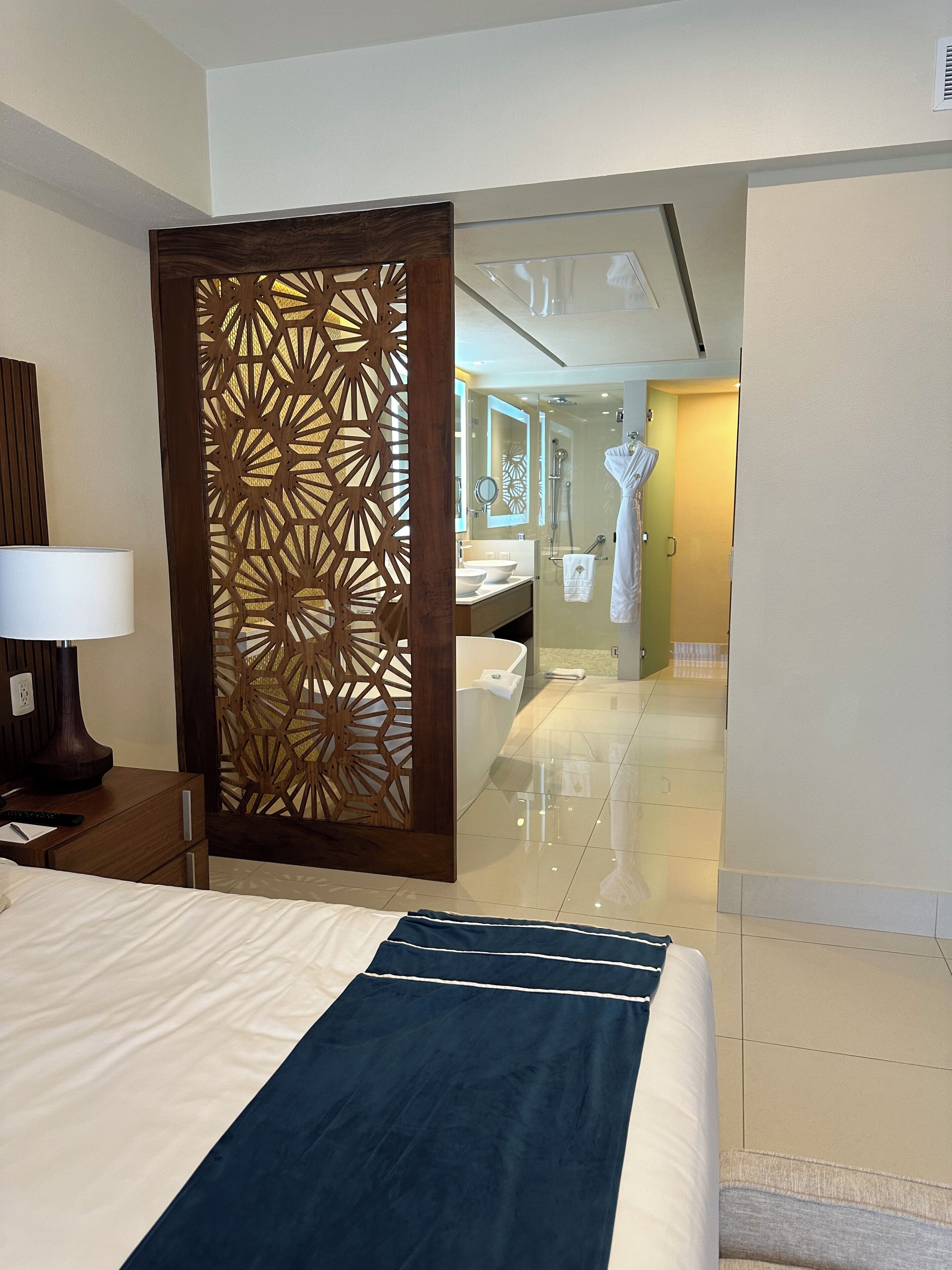 Habitación de hotel con cama, separador de madera, baño con puerta de vidrio y bata colgada en un perchero.