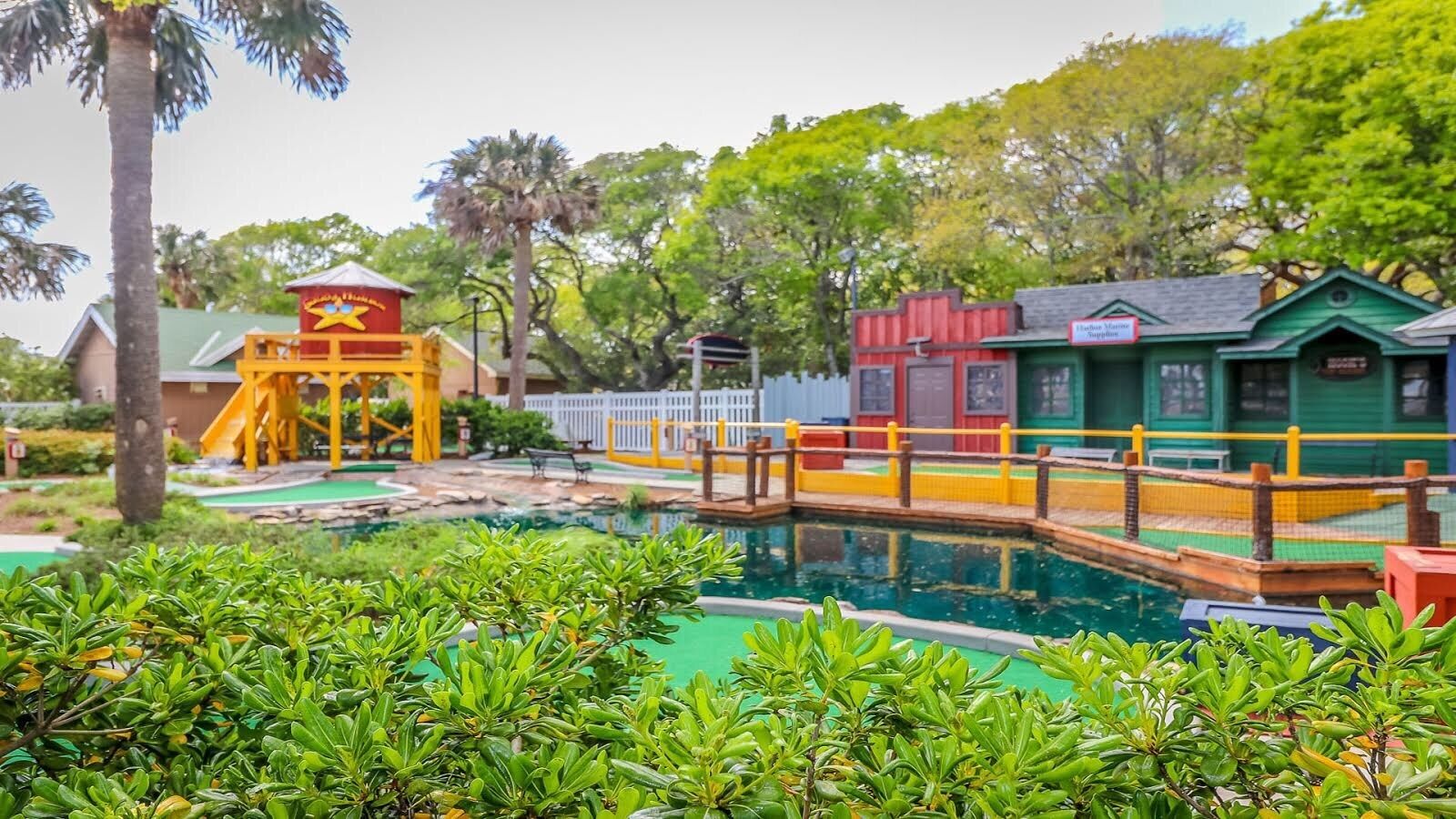 A pool area with a bridge, a yellow play structure, and a green building.